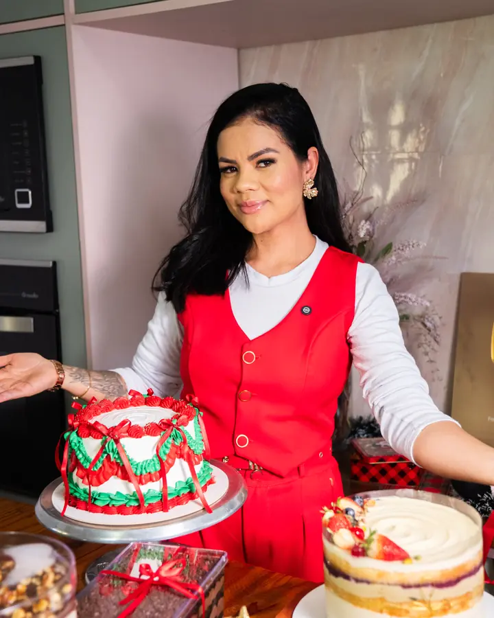 Karen Munhoz, confeiteira, de avental vermelho na cozinha, entre bolos decorados para o Natal